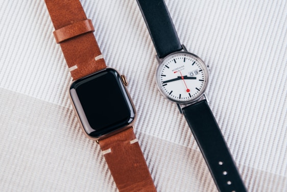 A modern digital smartwatch with a brown leather band is placed next to a classic analog wristwatch with a black leather band. The digital watch has a sleek, black rectangular face, while the analog watch features a white face with a date display and red accents on the hands.