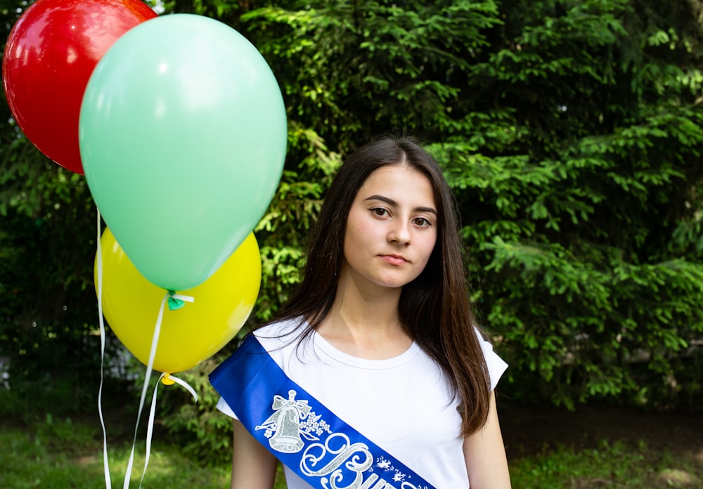 woman in white scoop neck t-shirt with blue sash holding three multicolored balloons