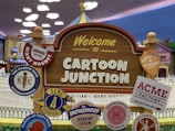 A playful outdoor sign welcomes visitors to 'Cartoon Junction.' It is surrounded by various whimsical badges featuring names like 'Order of the Giant Magnet,' 'The Karrot Club International,' and 'ACME Factory.' The background includes colorful cartoon-style buildings and a cloudy sky, enhancing the theme park ambiance.