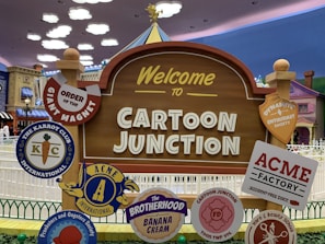 A playful outdoor sign welcomes visitors to 'Cartoon Junction.' It is surrounded by various whimsical badges featuring names like 'Order of the Giant Magnet,' 'The Karrot Club International,' and 'ACME Factory.' The background includes colorful cartoon-style buildings and a cloudy sky, enhancing the theme park ambiance.