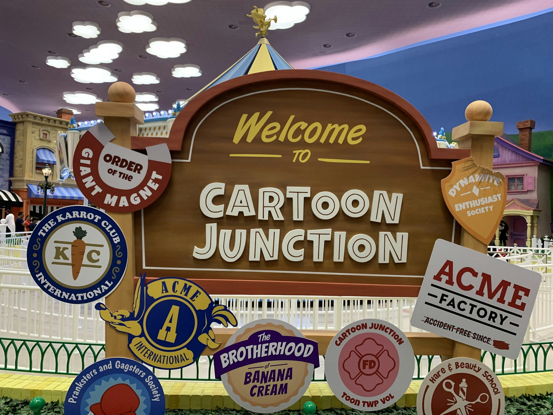 A playful outdoor sign welcomes visitors to 'Cartoon Junction.' It is surrounded by various whimsical badges featuring names like 'Order of the Giant Magnet,' 'The Karrot Club International,' and 'ACME Factory.' The background includes colorful cartoon-style buildings and a cloudy sky, enhancing the theme park ambiance.