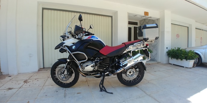 A large motorcycle is parked on a tiled pavement in front of a building with light-colored walls and closed garage doors. The motorcycle features a prominent red seat and is equipped with various accessories, including a windshield, a box at the rear, and side panniers. It is positioned on a kickstand, and to the side, there is a silver car and some green potted plants.