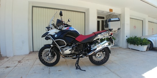 A large motorcycle is parked on a tiled pavement in front of a building with light-colored walls and closed garage doors. The motorcycle features a prominent red seat and is equipped with various accessories, including a windshield, a box at the rear, and side panniers. It is positioned on a kickstand, and to the side, there is a silver car and some green potted plants.
