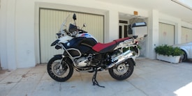 A large motorcycle is parked on a tiled pavement in front of a building with light-colored walls and closed garage doors. The motorcycle features a prominent red seat and is equipped with various accessories, including a windshield, a box at the rear, and side panniers. It is positioned on a kickstand, and to the side, there is a silver car and some green potted plants.