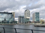 A series of modern office buildings, including one bearing the BBC logo, are positioned along a waterfront. The structures feature prominent glass facades, with varied architectural designs and heights. Overhead, the sky is overcast, casting a muted light on the scene.