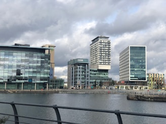 A series of modern office buildings, including one bearing the BBC logo, are positioned along a waterfront. The structures feature prominent glass facades, with varied architectural designs and heights. Overhead, the sky is overcast, casting a muted light on the scene.