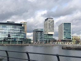 A series of modern office buildings, including one bearing the BBC logo, are positioned along a waterfront. The structures feature prominent glass facades, with varied architectural designs and heights. Overhead, the sky is overcast, casting a muted light on the scene.