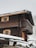 A wooden chalet-style house with a steeply pitched roof covered in snow. The structure has wooden window shutters painted in a light color, and a small balcony with intricate woodwork. Snow is falling gently, creating a wintry atmosphere.