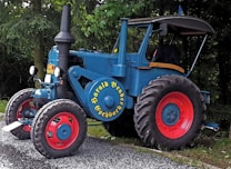 A vintage blue tractor with large red wheels is parked on a gravel path. The name 'Harald Bender Eschbach' is written in yellow on the side. It is surrounded by dense greenery, suggesting it is in a rural setting or forest area. The tractor has a large exhaust pipe and a covered driver's seat.