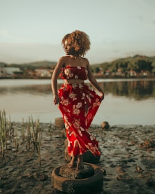A woman stands gracefully on a partially submerged tire in a mudflat. She is wearing a vibrant red dress with floral patterns, and her hair is styled in natural curls. The background features a serene body of water reflecting the cloudy sky, with hills and distant buildings lining the shore.