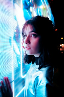 A young person with dark hair is gazing intently at a brightly illuminated and colorful digital screen. The scene is bathed in blue lighting, creating a futuristic and reflective atmosphere.