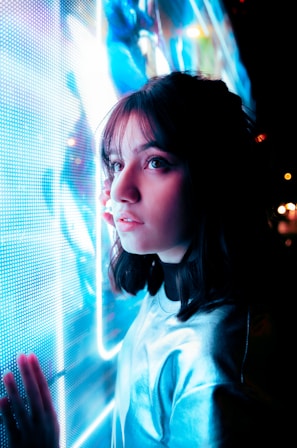 A young person with dark hair is gazing intently at a brightly illuminated and colorful digital screen. The scene is bathed in blue lighting, creating a futuristic and reflective atmosphere.