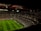 A large, illuminated stadium filled with a vast audience. The field is marked for soccer, with players actively participating in a match. The crowd is engaged, with numerous lights visible in the stands, possibly from phones. The stadium's roof structure is visible with red beams, and an electronic scoreboard shows a score of 0-0.