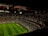 A large, illuminated stadium filled with a vast audience. The field is marked for soccer, with players actively participating in a match. The crowd is engaged, with numerous lights visible in the stands, possibly from phones. The stadium's roof structure is visible with red beams, and an electronic scoreboard shows a score of 0-0.