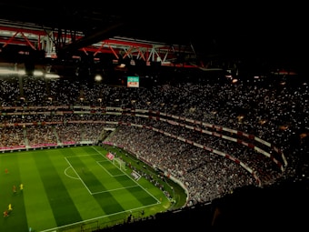 A large, illuminated stadium filled with a vast audience. The field is marked for soccer, with players actively participating in a match. The crowd is engaged, with numerous lights visible in the stands, possibly from phones. The stadium's roof structure is visible with red beams, and an electronic scoreboard shows a score of 0-0.