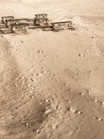 Several wooden picnic tables are scattered across a sandy beach with numerous footprints throughout the area.