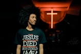 A person with curly hair is wearing a black t-shirt with the text 'John 10:10 JESUS DIED SO I COULD LIVE' in a dimly lit environment. In the background, there is a prominent glowing red cross on the wall.