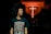 A person with curly hair is wearing a black t-shirt with the text 'John 10:10 JESUS DIED SO I COULD LIVE' in a dimly lit environment. In the background, there is a prominent glowing red cross on the wall.