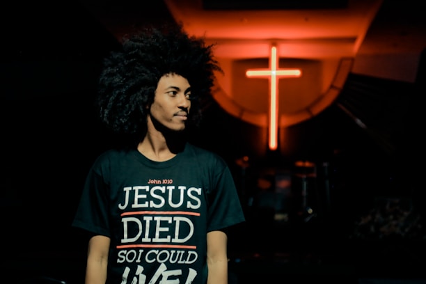 A person with curly hair is wearing a black t-shirt with the text 'John 10:10 JESUS DIED SO I COULD LIVE' in a dimly lit environment. In the background, there is a prominent glowing red cross on the wall.