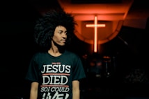 A person with curly hair is wearing a black t-shirt with the text 'John 10:10 JESUS DIED SO I COULD LIVE' in a dimly lit environment. In the background, there is a prominent glowing red cross on the wall.