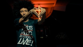 A person with a large afro hairstyle stands in a dimly lit environment. They are making a heart shape with their hands in front of a light source that creates a warm, orange glow in the background. The person is wearing a dark t-shirt with white and red text.