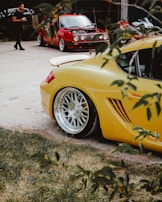 A vibrant scene with a yellow sports car prominently in the foreground, showcasing its sleek design and polished wheels. In the background, there's a red classic car with its hood open, possibly being attended to or presented. A person is walking nearby, holding a camera, suggesting an informal car meet or photography session. The view is partially framed by branches and leaves, adding a natural element to the urban setting.