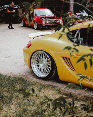 A vibrant scene with a yellow sports car prominently in the foreground, showcasing its sleek design and polished wheels. In the background, there's a red classic car with its hood open, possibly being attended to or presented. A person is walking nearby, holding a camera, suggesting an informal car meet or photography session. The view is partially framed by branches and leaves, adding a natural element to the urban setting.