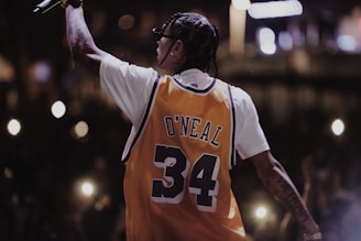 A person with braided hair is wearing a yellow basketball jersey with the name 'O'Neal' and the number '34'. They are holding a microphone and facing away from the camera, suggesting a performance or concert setting. The background is dark with illuminated spots from camera flashes or lights, indicating an audience.