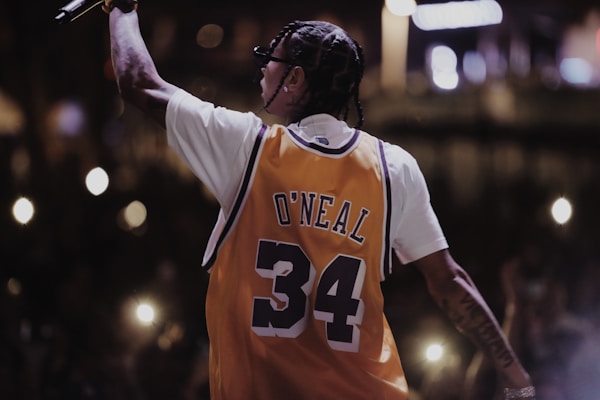 A person with braided hair is wearing a yellow basketball jersey with the name 'O'Neal' and the number '34'. They are holding a microphone and facing away from the camera, suggesting a performance or concert setting. The background is dark with illuminated spots from camera flashes or lights, indicating an audience.