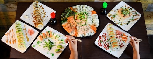 A variety of sushi dishes arranged on a dark wooden table. Several plates display different types of sushi rolls, garnished with sauces, avocado, fish, and greenery. In the center is a large platter with sushi rolls featuring salmon, tuna, and cucumber. Two small bottles of soy sauce, one with a green cap and one with a red cap, are present. Two pairs of chopsticks are actively picking up pieces of sushi from the various plates.