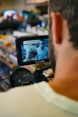 A person operating a camera, focusing on a small monitor displaying an image of two people in a store setting. The background is blurred, showing shelves with various items, creating a busy atmosphere.