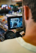 A person operating a camera, focusing on a small monitor displaying an image of two people in a store setting. The background is blurred, showing shelves with various items, creating a busy atmosphere.
