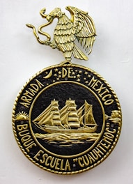 A decorative emblem features a golden eagle with outstretched wings perched on a circular crest. Inside the crest, a detailed gold depiction of a sailing ship with three masts is surrounded by stars and celestial symbols. The text around the circle reads 'ARMADA DE MEXICO BUQUE ESCUELA 'CUAUHTEMOC'.'