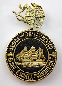A decorative emblem features a golden eagle with outstretched wings perched on a circular crest. Inside the crest, a detailed gold depiction of a sailing ship with three masts is surrounded by stars and celestial symbols. The text around the circle reads 'ARMADA DE MEXICO BUQUE ESCUELA 'CUAUHTEMOC'.'