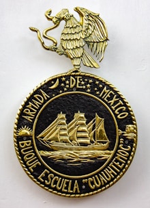 A decorative emblem features a golden eagle with outstretched wings perched on a circular crest. Inside the crest, a detailed gold depiction of a sailing ship with three masts is surrounded by stars and celestial symbols. The text around the circle reads 'ARMADA DE MEXICO BUQUE ESCUELA 'CUAUHTEMOC'.'
