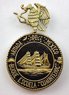 A decorative emblem features a golden eagle with outstretched wings perched on a circular crest. Inside the crest, a detailed gold depiction of a sailing ship with three masts is surrounded by stars and celestial symbols. The text around the circle reads 'ARMADA DE MEXICO BUQUE ESCUELA 'CUAUHTEMOC'.'