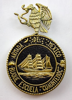 A decorative emblem features a golden eagle with outstretched wings perched on a circular crest. Inside the crest, a detailed gold depiction of a sailing ship with three masts is surrounded by stars and celestial symbols. The text around the circle reads 'ARMADA DE MEXICO BUQUE ESCUELA 'CUAUHTEMOC'.'