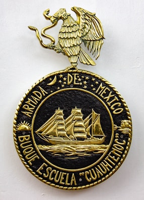 A decorative emblem features a golden eagle with outstretched wings perched on a circular crest. Inside the crest, a detailed gold depiction of a sailing ship with three masts is surrounded by stars and celestial symbols. The text around the circle reads 'ARMADA DE MEXICO BUQUE ESCUELA 'CUAUHTEMOC'.'