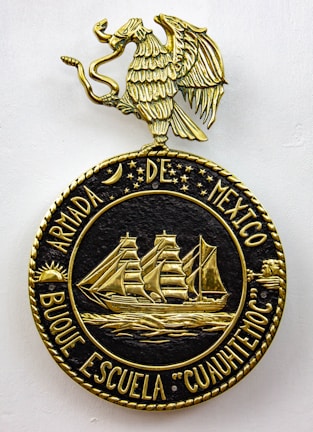 A decorative emblem features a golden eagle with outstretched wings perched on a circular crest. Inside the crest, a detailed gold depiction of a sailing ship with three masts is surrounded by stars and celestial symbols. The text around the circle reads 'ARMADA DE MEXICO BUQUE ESCUELA 'CUAUHTEMOC'.'
