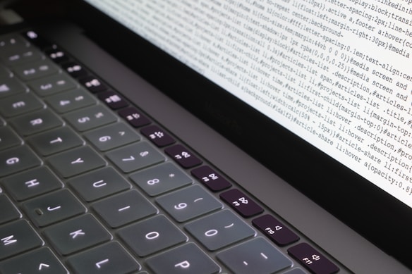 A close-up view of a partially illuminated laptop keyboard. The screen displays lines of code or text in a programming or command-line environment. The keys are dark with bright white lettering, and the text on the screen appears densely packed.