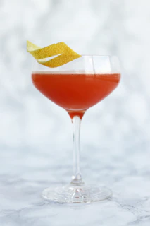 An elegant cocktail glass filled with a vibrant red drink, garnished with a twist of orange peel against a dark background.