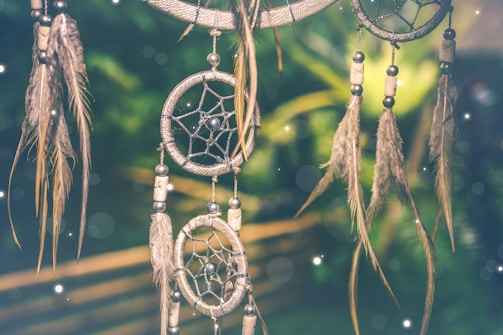 A dreamcatcher with intricate webbing and adorned with feathers and beads, set against a blurred green background with soft glowing orbs of light.