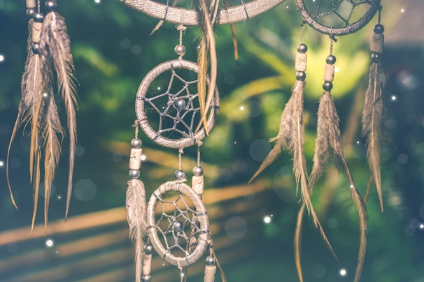 A dreamcatcher with intricate webbing and adorned with feathers and beads, set against a blurred green background with soft glowing orbs of light.