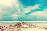 A beach scene with a large pile of debris, including driftwood and plastic waste, in the foreground. The ocean waves gently crash in the background under a partly cloudy sky. Several people can be seen in the distance, enjoying the water.
