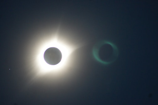 A solar eclipse with the moon obscuring the sun, creating a bright halo effect. The surrounding sky is dark, and an additional lens flare effect is visible to the right of the eclipse.