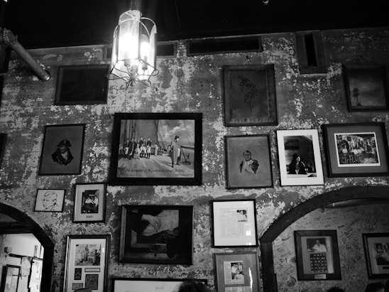 Multiple framed pictures and artworks hang on a textured, worn wall in an eclectic arrangement. A prominent light fixture hangs from the ceiling, casting a glow over the artwork, which includes portraits, scene depictions, and abstract pieces.