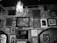 Multiple framed pictures and artworks hang on a textured, worn wall in an eclectic arrangement. A prominent light fixture hangs from the ceiling, casting a glow over the artwork, which includes portraits, scene depictions, and abstract pieces.