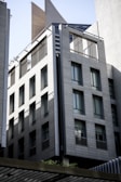 A modern multi-story building with a sleek design features large rectangular windows and a facade made of light gray stone or concrete panels. The building is in an urban setting and includes architectural details like thin steel beams and sunshades.