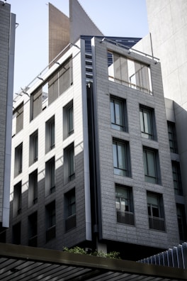 A modern multi-story building with a sleek design features large rectangular windows and a facade made of light gray stone or concrete panels. The building is in an urban setting and includes architectural details like thin steel beams and sunshades.