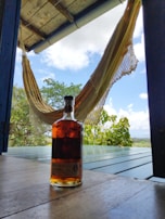 A bottle of rum is placed on a wooden porch floor with a relaxed hammock hanging in the background. The scene extends to a lush green landscape with a clear blue sky dotted with clouds.